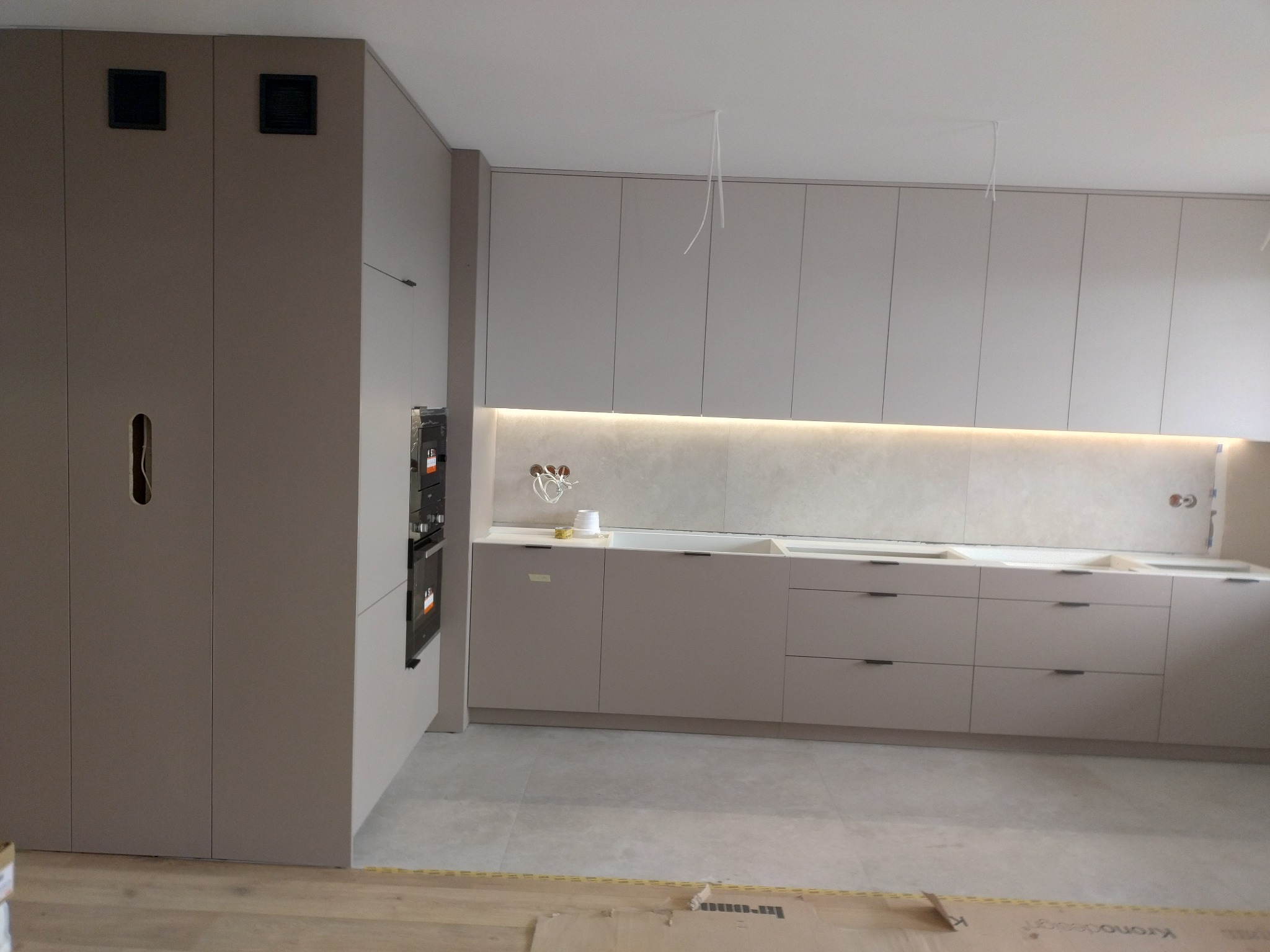 Kuchnia w trakcie montażu: beżowe szafki dolne z ciemnymi uchwytami i podświetleniem LED, wysoka zabudowa z piekarnikiem, widoczne kable i kartony na podłodze.