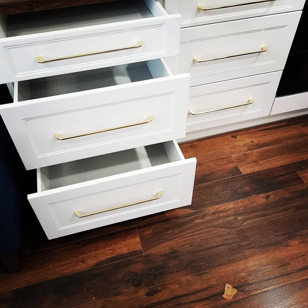 Białe szuflady z frezowanymi frontami i złotymi uchwytami, dwie otwarte, na tle ciemnej, drewnianej podłogi.