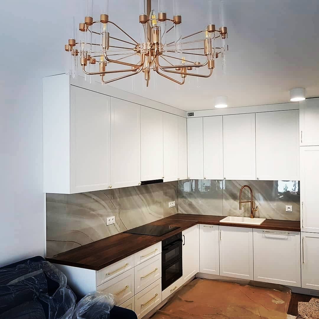 Biała kuchnia z ciemnym drewnianym blatem, złotymi uchwytami i marmurowym backsplashiem, oświetlona ozdobnym, złotym żyrandolem