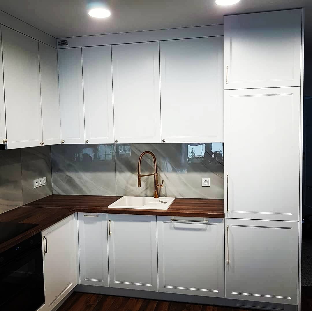 Jasna kuchnia z białymi frontami szafek, drewnianym blatem i marmurowym panelem nad blatem, bateria w kolorze miedzi.