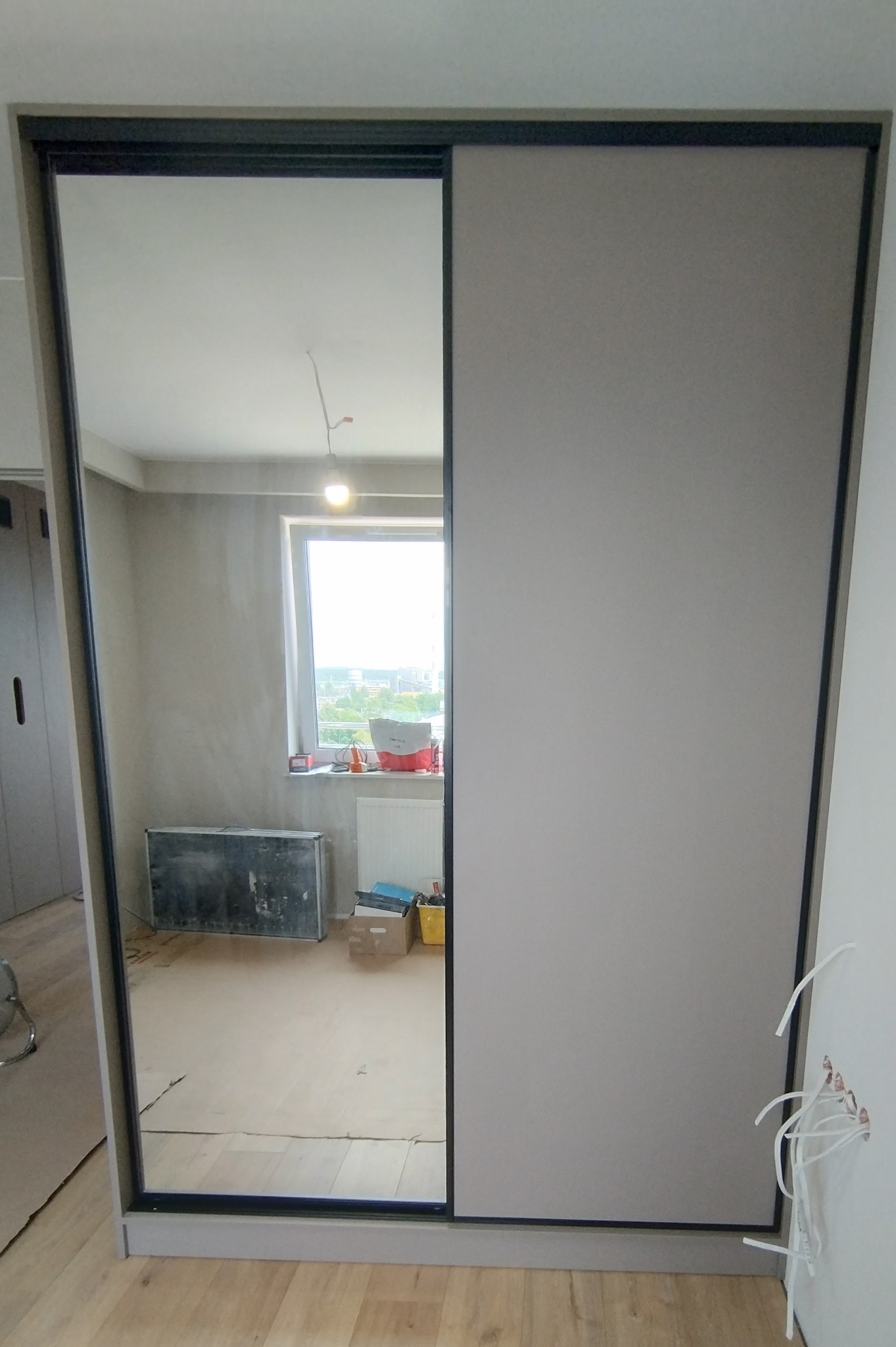 Szafa przesuwna z lustrem w trakcie montażu, widoczne kable wystające ze ściany i odblask pokoju z materiałami budowlanymi.