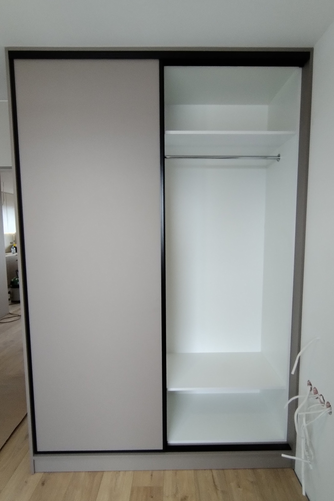 Nowoczesna szafa wnękowa z przesuwnymi drzwiami, częściowo otwarta, z widocznymi półkami i drążkiem na ubrania, surowe wykończenie wnętrza z wystającymi kablami.