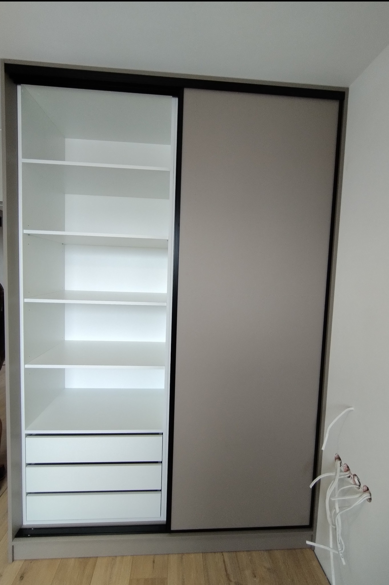 Szafa wnękowa z przesuwnymi drzwiami, widoczna otwarta część z białymi półkami i trzema szufladami, obok ściana z wystającymi kablami elektrycznymi.