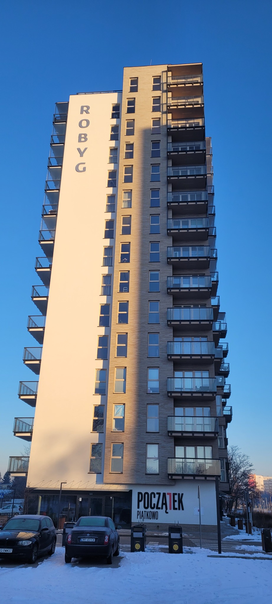 Wysoki budynek mieszkalny z balkonami, częściowo oświetlony słońcem, z napisem 'ROBYG' na bocznej ścianie i widocznymi samochodami na parkingu pokrytym śniegiem.