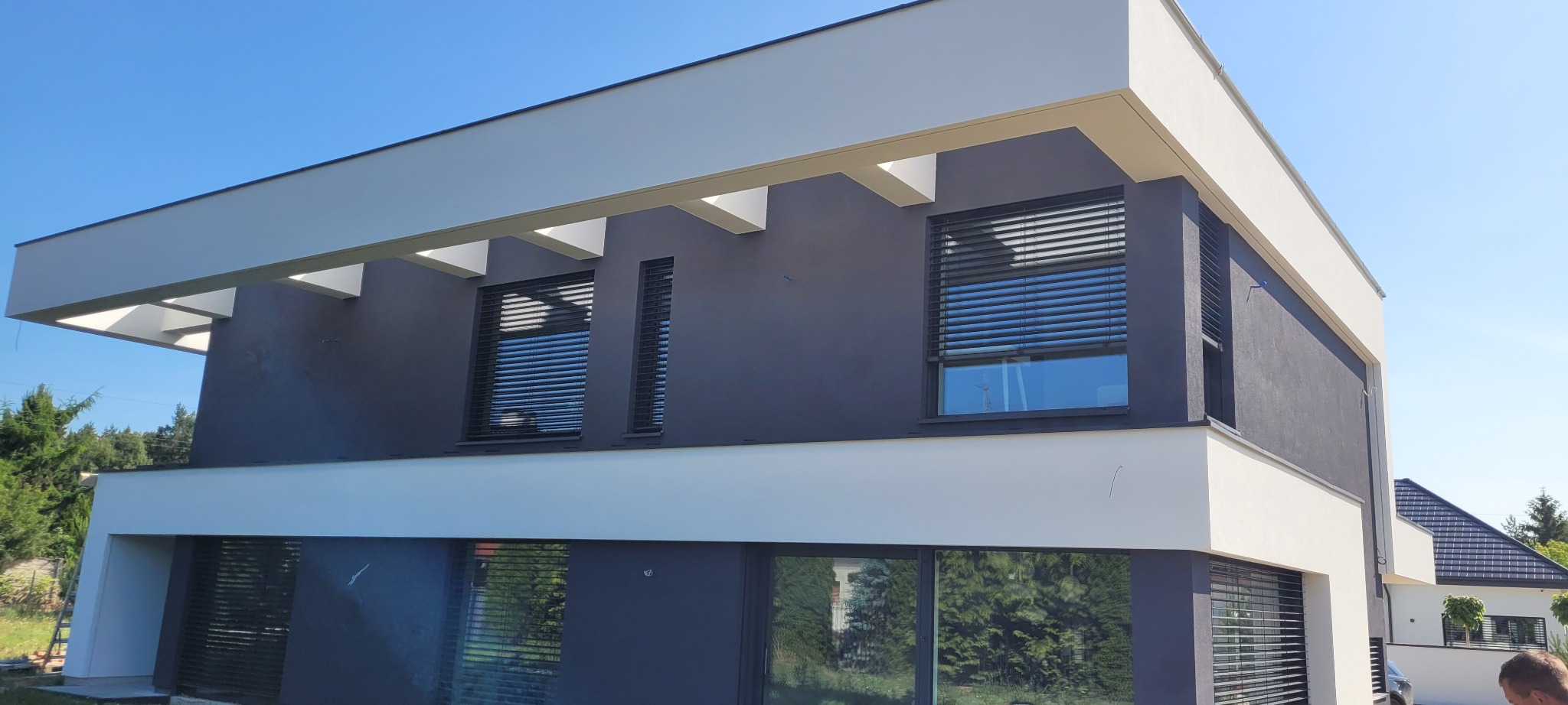 Dwukondygnacyjny dom z grafitową elewacją, białymi elementami architektonicznymi i żaluzjami zewnętrznymi w oknach, widziany w słoneczny dzień.