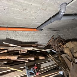 Hydro-Raf - Szare i pomarańczowe rury podwieszone pod sufitem w piwnicy z ceglanymi ścianami. Na dole widoczne narzędzia i stos drewna.