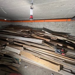 Hydro-Raf - Pomarańczowa rura kanalizacyjna pod betonowym sufitem piwnicy, oświetlona lampą roboczą. Na podłodze sterta drewna i wiertarka akumulatorowa.