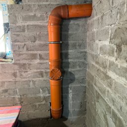 Hydro-Raf - Pomarańczowa rura kanalizacyjna z obejmami przymocowana do szarej, surowej ściany z cegieł. Rura wchodzi w ścianę i podłoże. Widoczny fragment okna i podłoża.