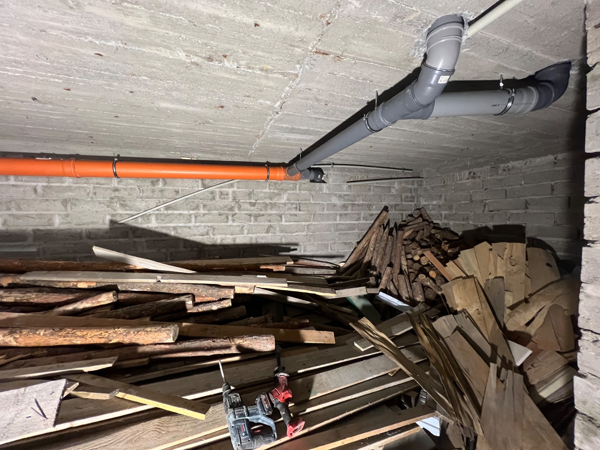 Szare i pomarańczowe rury podwieszone pod sufitem w piwnicy z ceglanymi ścianami. Na dole widoczne narzędzia i stos drewna.