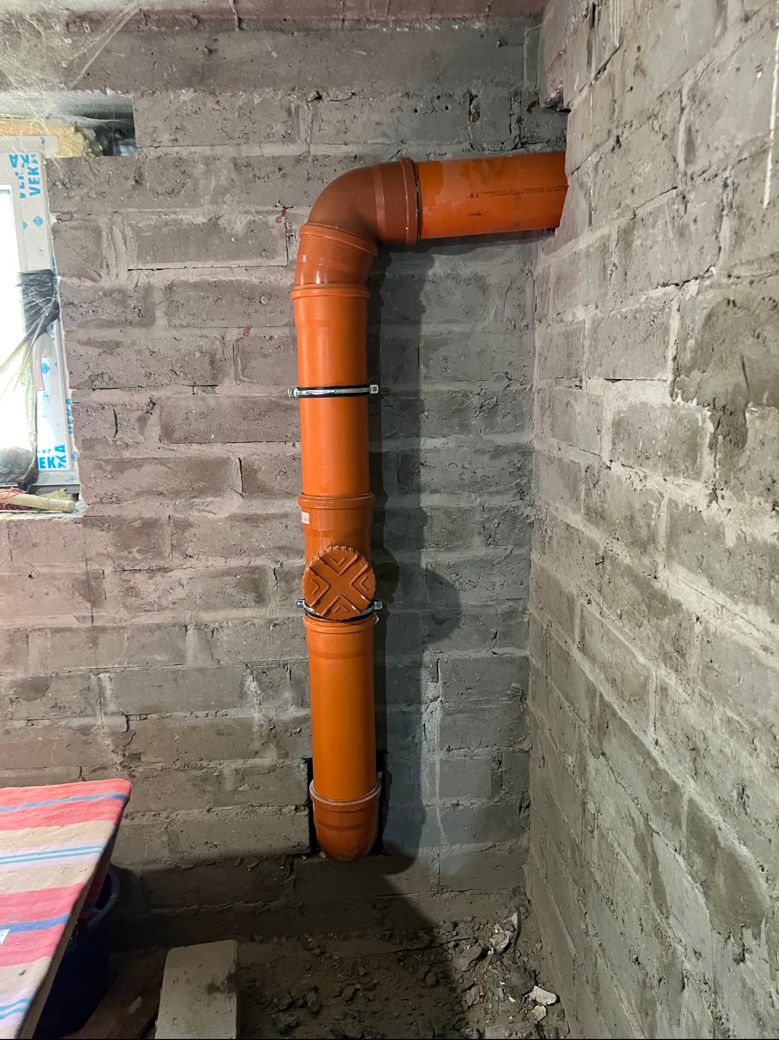 Pomarańczowa rura kanalizacyjna z obejmami przymocowana do szarej, surowej ściany z cegieł. Rura wchodzi w ścianę i podłoże. Widoczny fragment okna i podłoża.