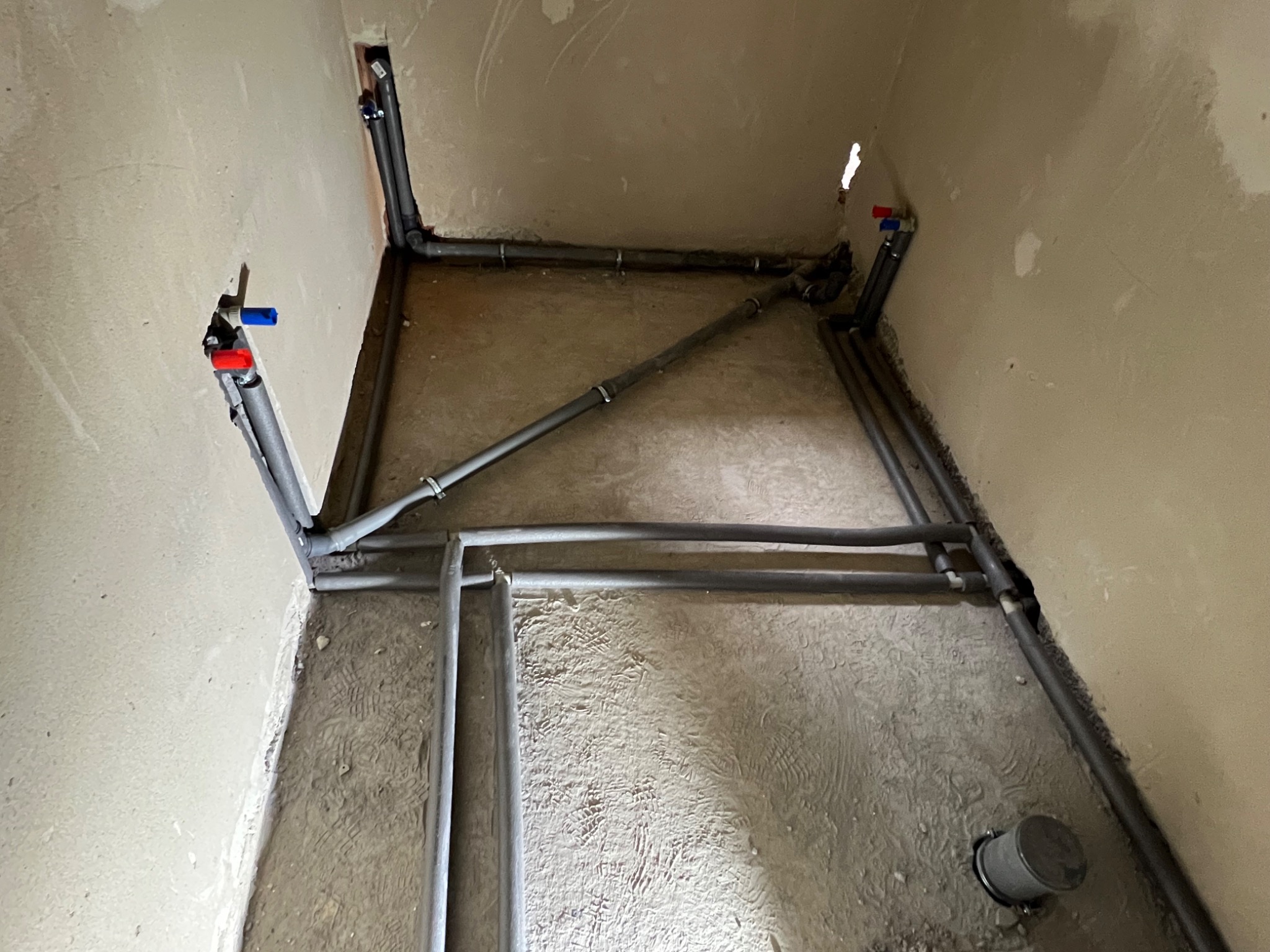 Szare rury instalacji wodnej ułożone w surowym pomieszczeniu z widocznymi przyłączami do armatury. Etap prac instalacyjnych w nowym budynku.