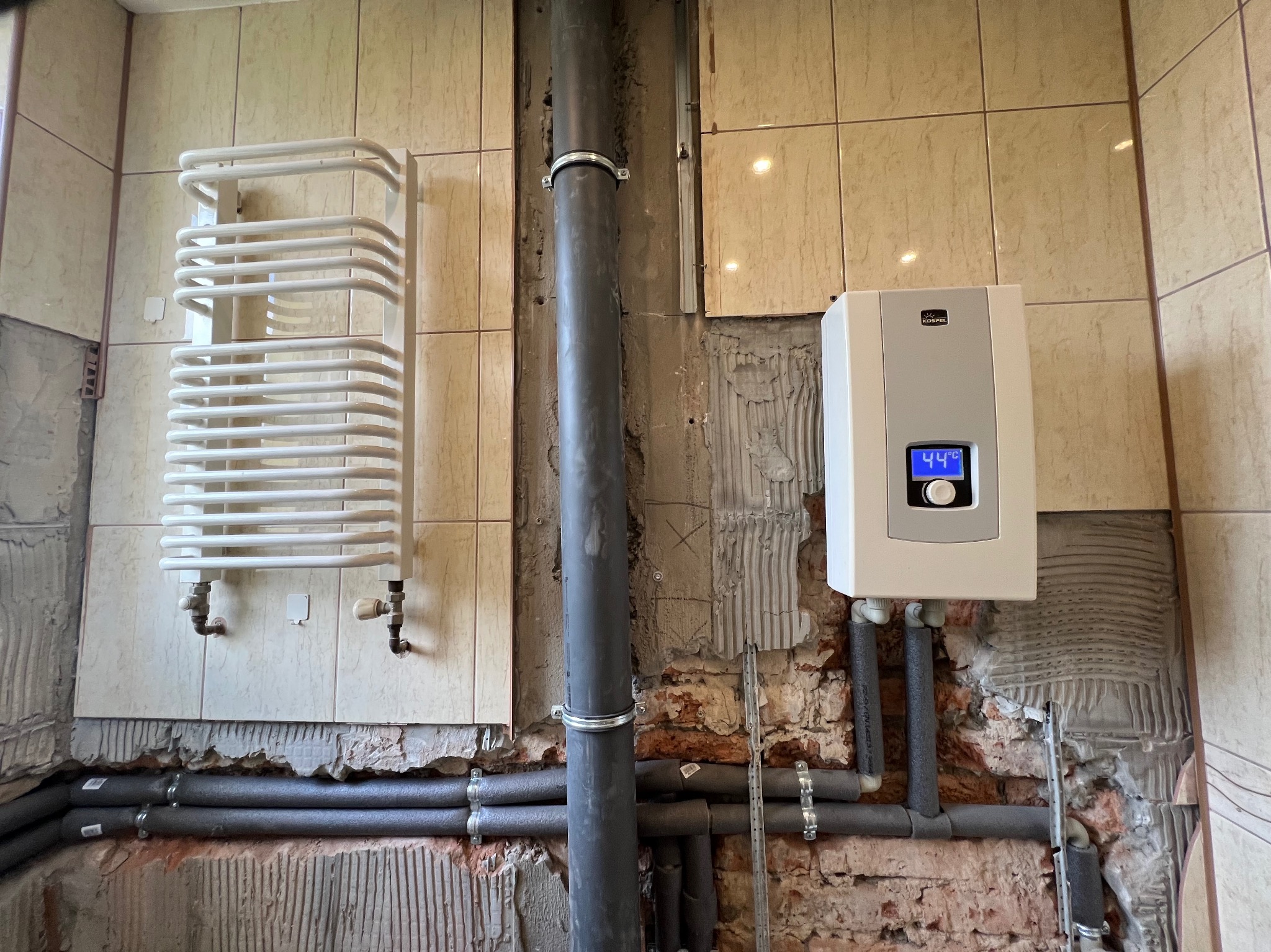 Biały grzejnik drabinkowy, podgrzewacz wody Kospel z wyświetlaczem 44°C i rury w izolacji, na tle remontowanej łazienki z beżowymi płytkami i odsłoniętą cegłą.