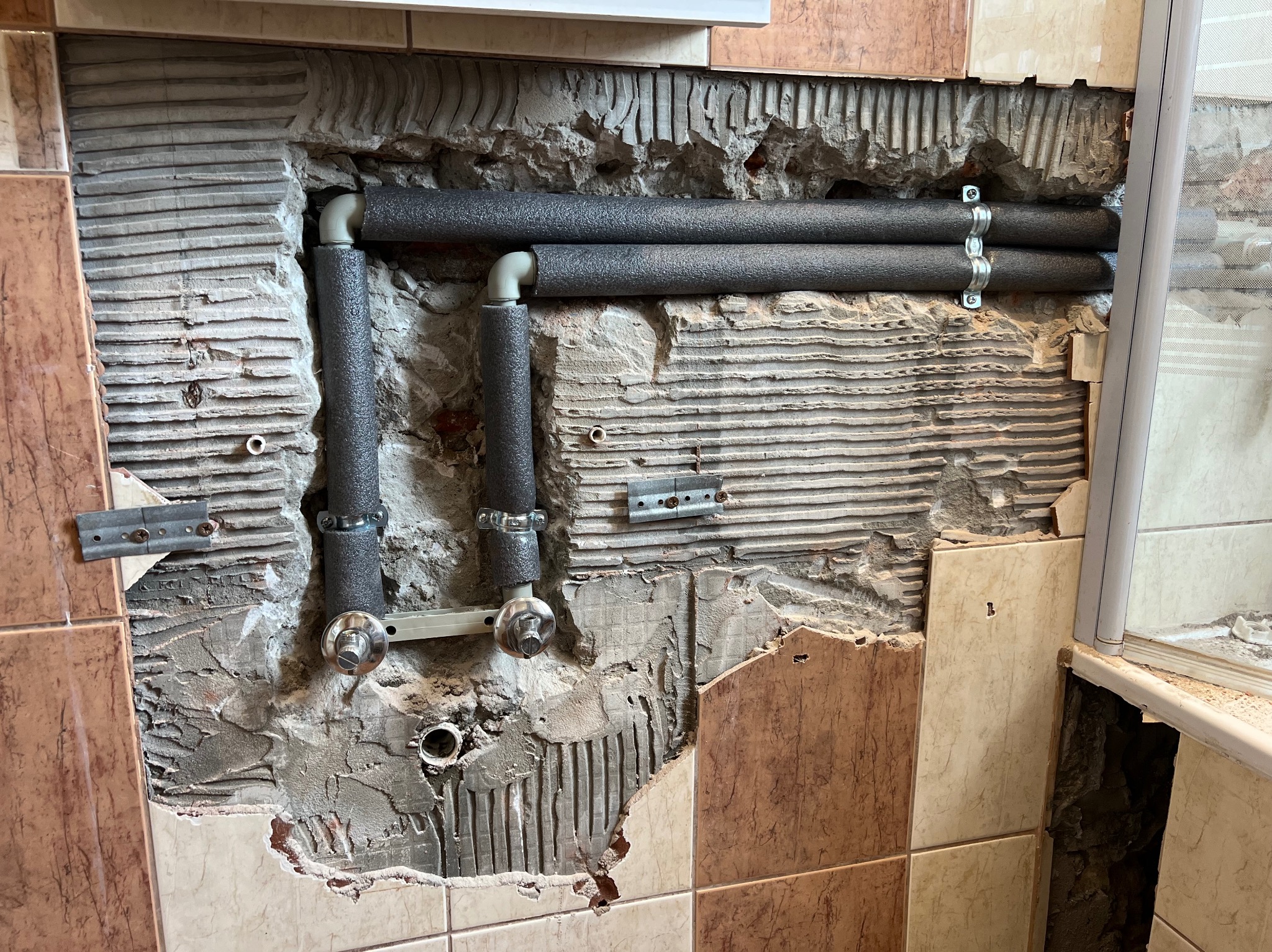 Odsłonięte rury w izolacji termicznej, częściowo zamontowane na ścianie z uszkodzonymi płytkami. Widoczne elementy mocujące i fragment instalacji w łazience.