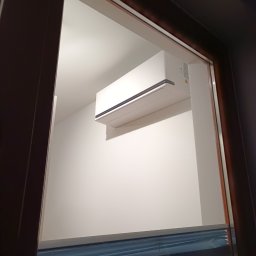 MEJK 4 U SPÓŁKA Z OGRANICZONĄ ODPOWIEDZIALNOŚCIĄ - Biała jednostka klimatyzacji zamontowana na ścianie w pokoju, widoczna przez okno z częściowo zasłoniętymi żaluzjami.
