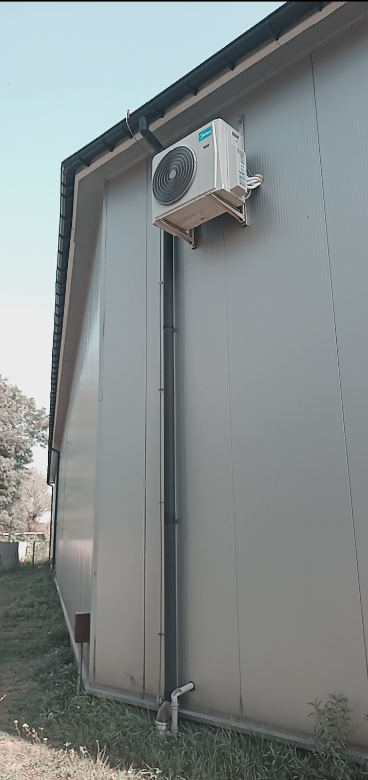 Boczny widok na zamontowaną jednostkę zewnętrzną klimatyzatora Midea na ścianie budynku z blachy trapezowej, z widocznymi rurami odprowadzającymi skropliny.