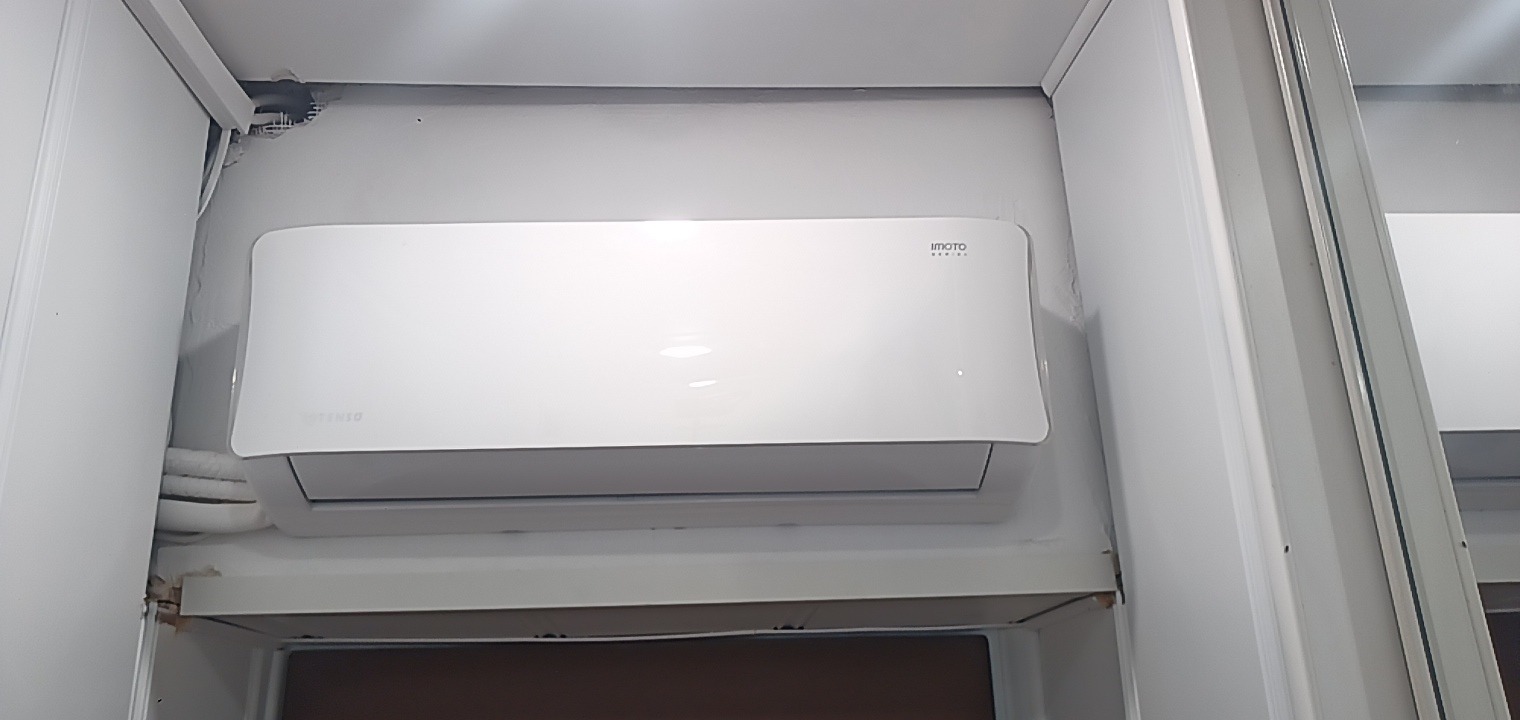 Biała jednostka wewnętrzna klimatyzatora marki Imoto zamontowana w rogu pomieszczenia, widoczne rury instalacyjne i fragment zabudowy.