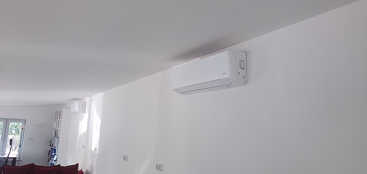 Biały klimatyzator ścienny zamontowany w minimalistycznym, jasnym wnętrzu z widocznym fragmentem regału z książkami i czerwonej sofy w tle.