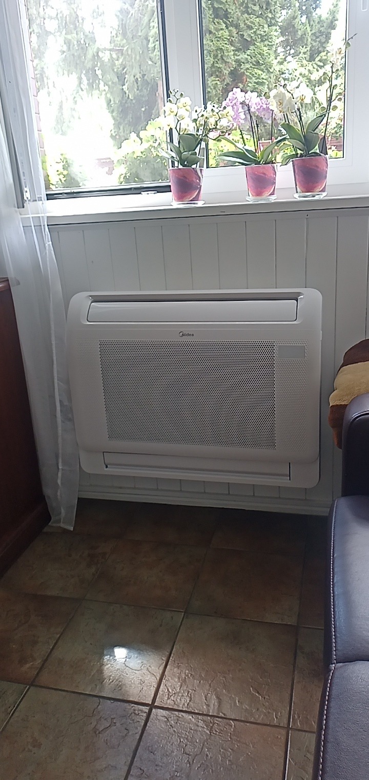 Biała jednostka klimatyzacji Midea zamontowana przy ścianie pod oknem z trzema doniczkami z białymi i fioletowymi orchideami, widok na zielone drzewa za oknem, brązowe płytki podłogowe.