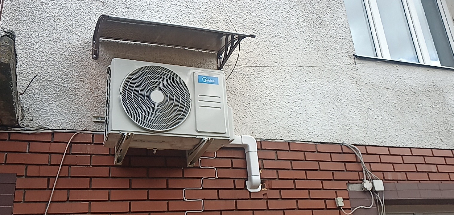 Zewnętrzna jednostka klimatyzacji Midea zamontowana na ścianie budynku z cegły, widoczne przewody i rurki odprowadzające skropliny, nad jednostką daszek ochronny.