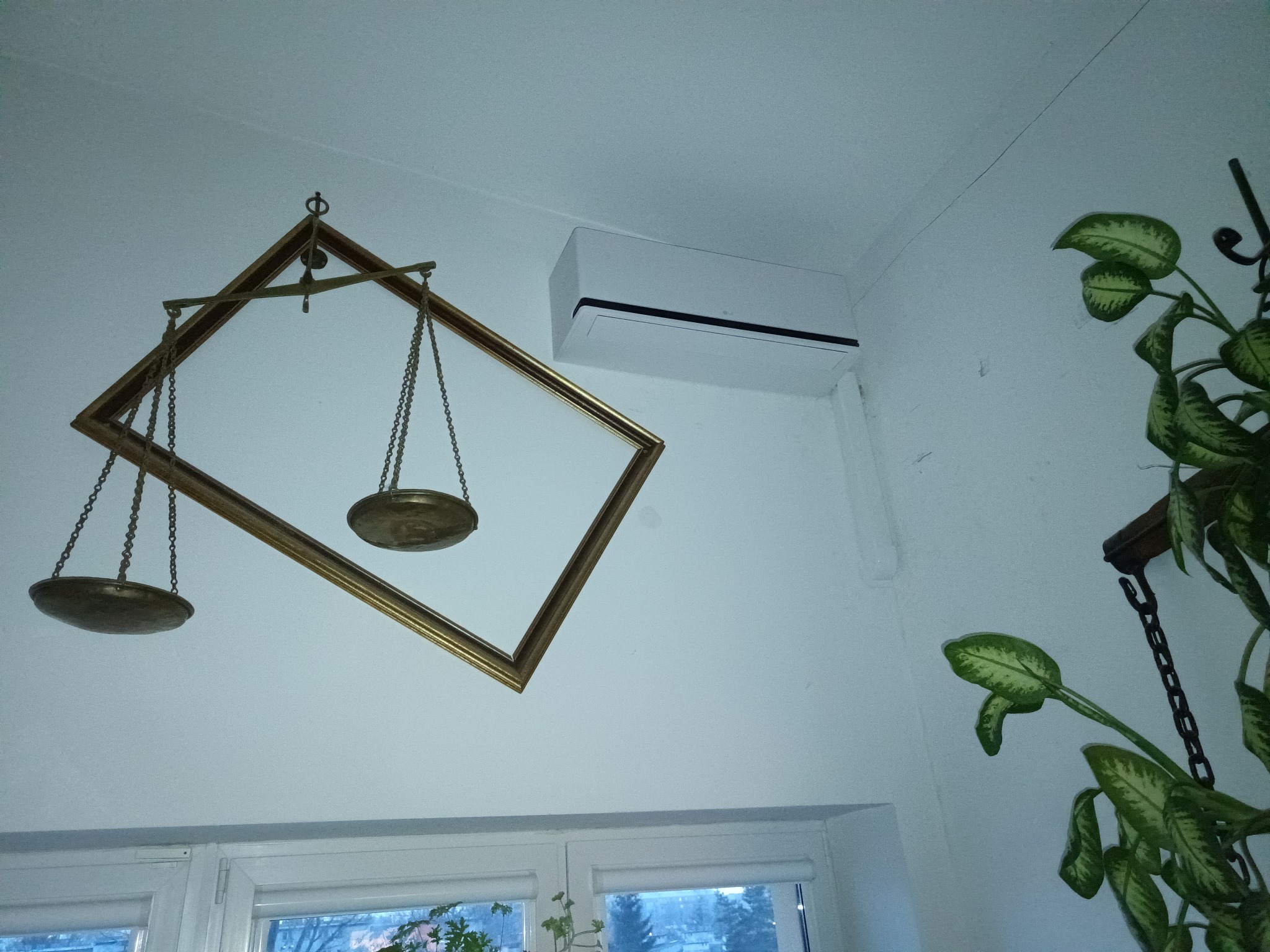 Biała jednostka klimatyzacyjna na ścianie w pomieszczeniu z dekoracyjnymi, złotymi wagami wiszącymi pod sufitem i rośliną doniczkową w rogu.