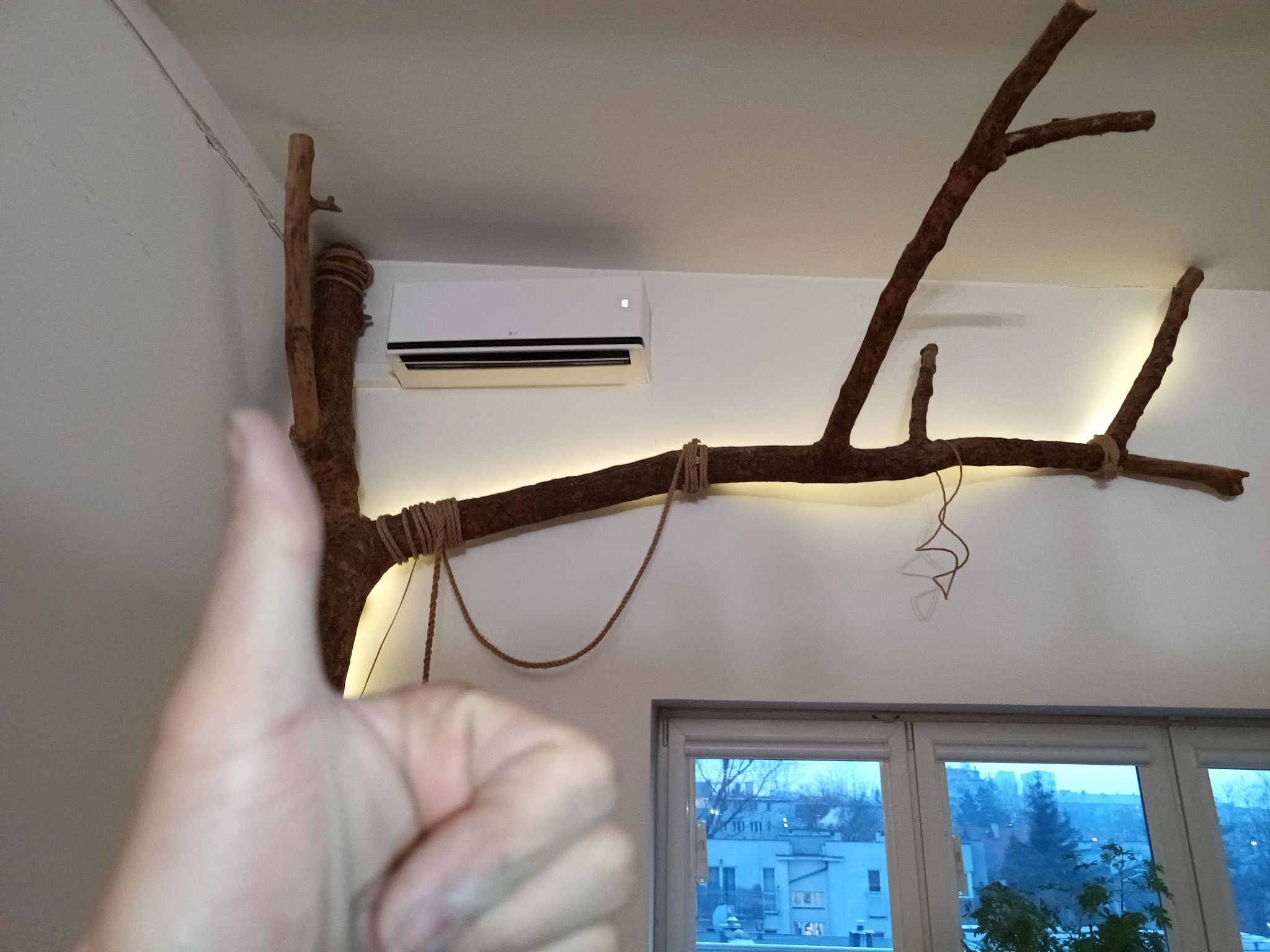 Biały klimatyzator LG zamontowany na ścianie, udekorowany drewnianą gałęzią z oświetleniem LED, przymocowaną sznurkiem, widok z kciukiem w górę.