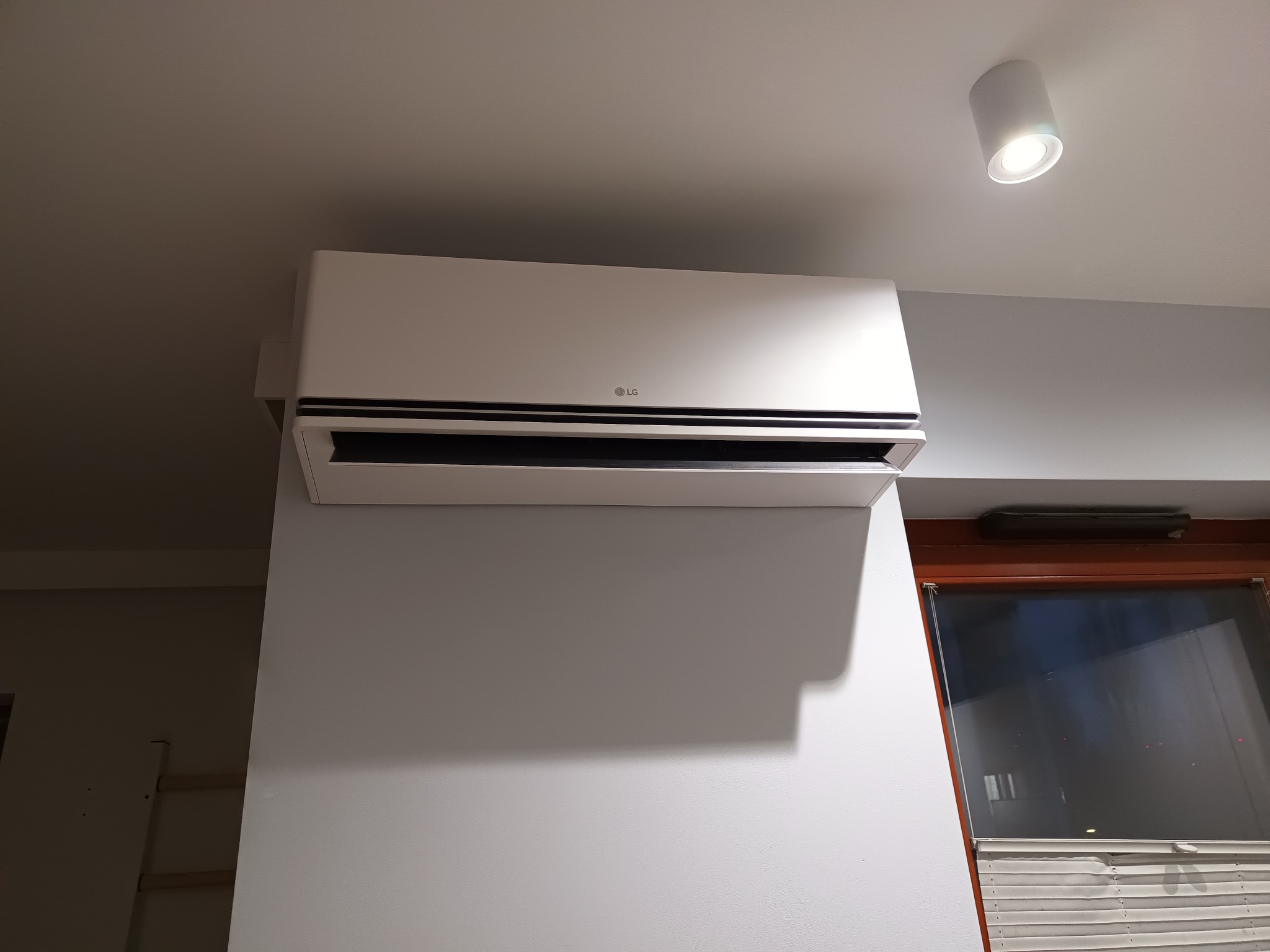 Biała jednostka wewnętrzna klimatyzacji LG zamontowana na szarej ścianie, widoczna lampa sufitowa i fragment okna w tle.