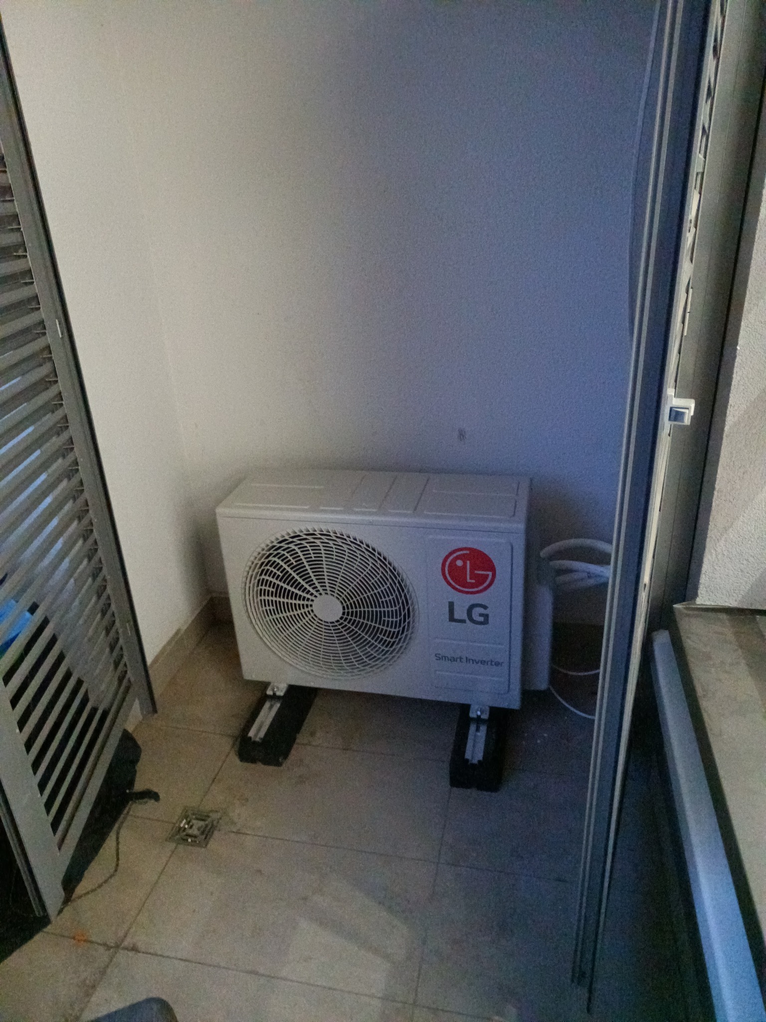 Biała jednostka zewnętrzna klimatyzacji LG Smart Inverter, zamontowana na czarnych wspornikach w narożniku balkonu, widoczne rury i kratka ściekowa w podłodze.