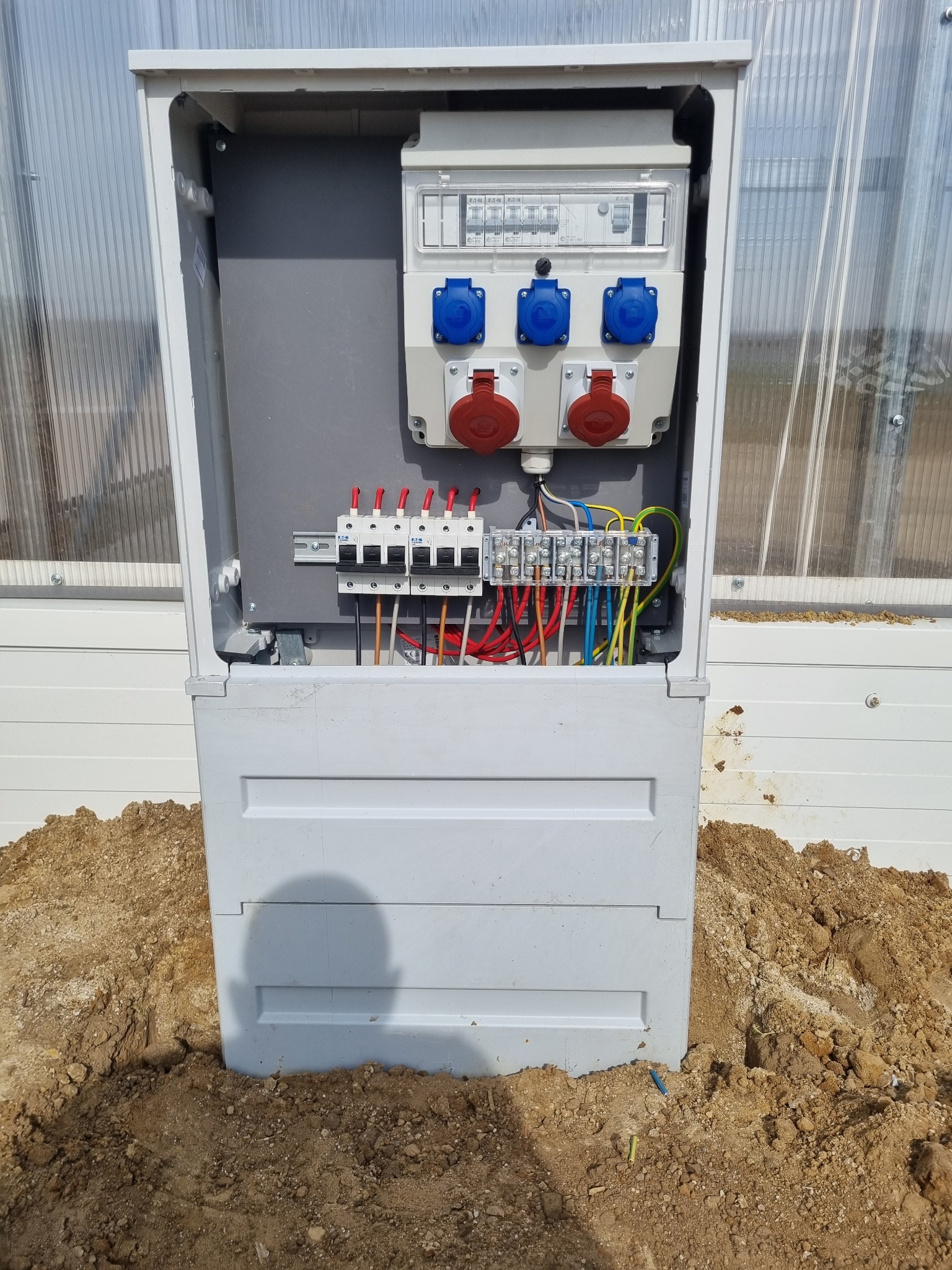 Otwarta skrzynka elektryczna z widocznymi bezpiecznikami, gniazdami siłowymi (czerwone i niebieskie) i przewodami, umieszczona na zewnątrz budynku z przezroczystymi ścianami, otoczona ziemią.