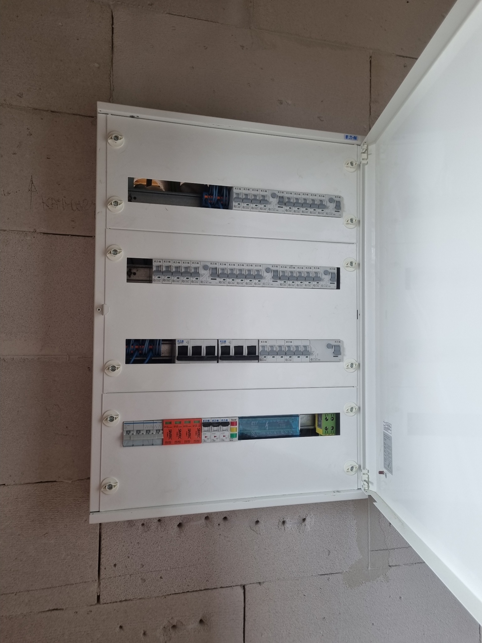 Otwarta skrzynka rozdzielcza z bezpiecznikami i przewodami zamontowana na surowej ścianie z betonu komórkowego, widoczne zabezpieczenia Eaton