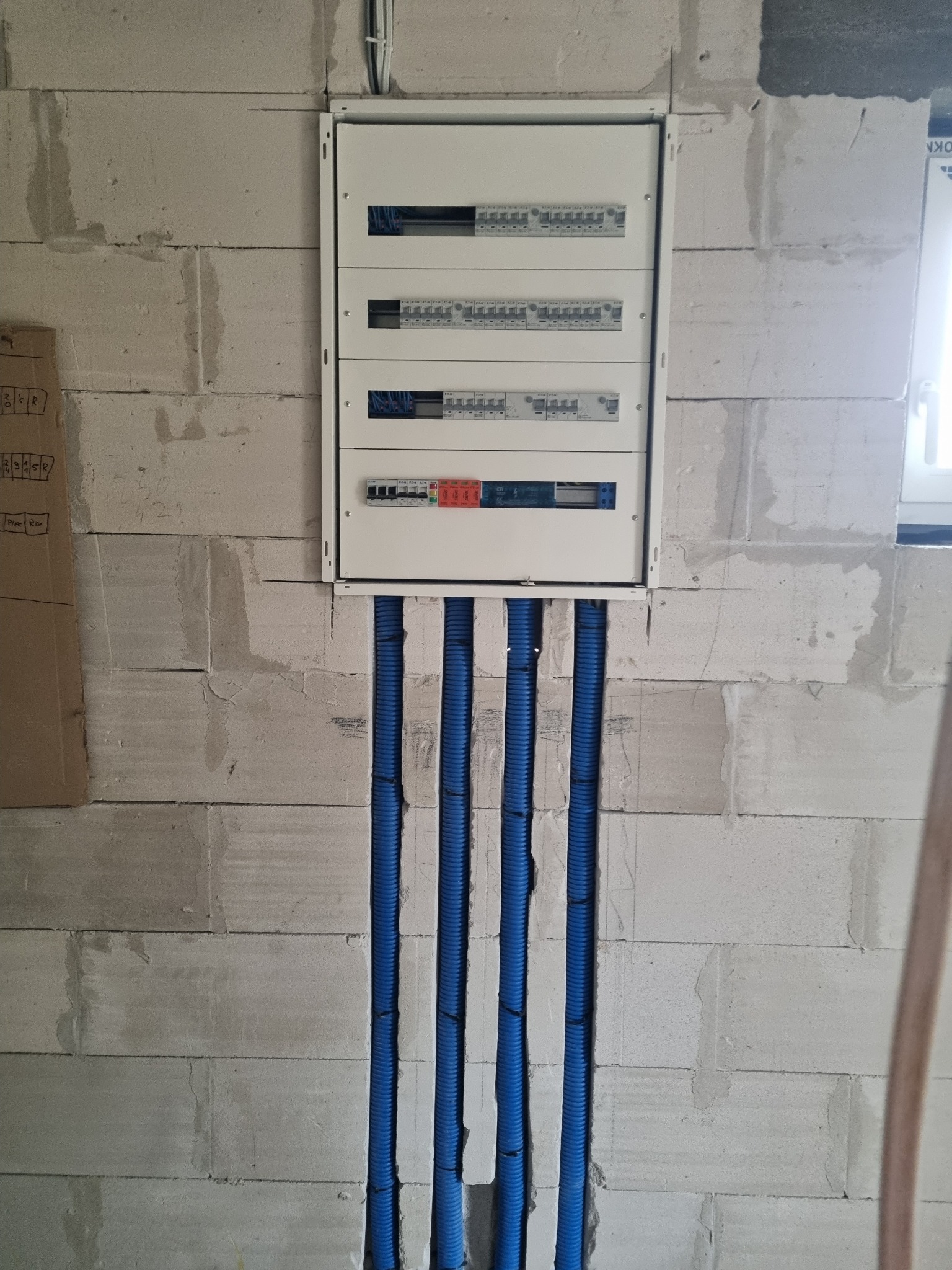 Rozdzielnia elektryczna z czterema rzędami zabezpieczeń, zamontowana na surowej ścianie z betonu komórkowego, z niebieskimi rurami karbowanymi prowadzącymi od dołu.