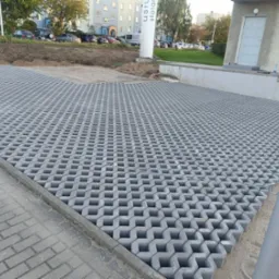 Ułożona szara ażurowa kostka brukowa na przygotowanym terenie, widoczna krawędź chodnika z kostki brukowej i budynek w tle.