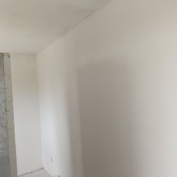 Radosław Jasiński - Świeżo pomalowane na biało ściany w rogu pokoju, z widocznym surowym wejściem do innego pomieszczenia z lewej strony, ukazujące etap prac wykończeniowych.