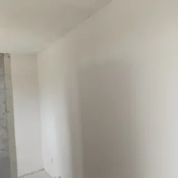 Świeżo pomalowane na biało ściany w rogu pokoju, z widocznym surowym wejściem do innego pomieszczenia z lewej strony, ukazujące etap prac wykończeniowych.