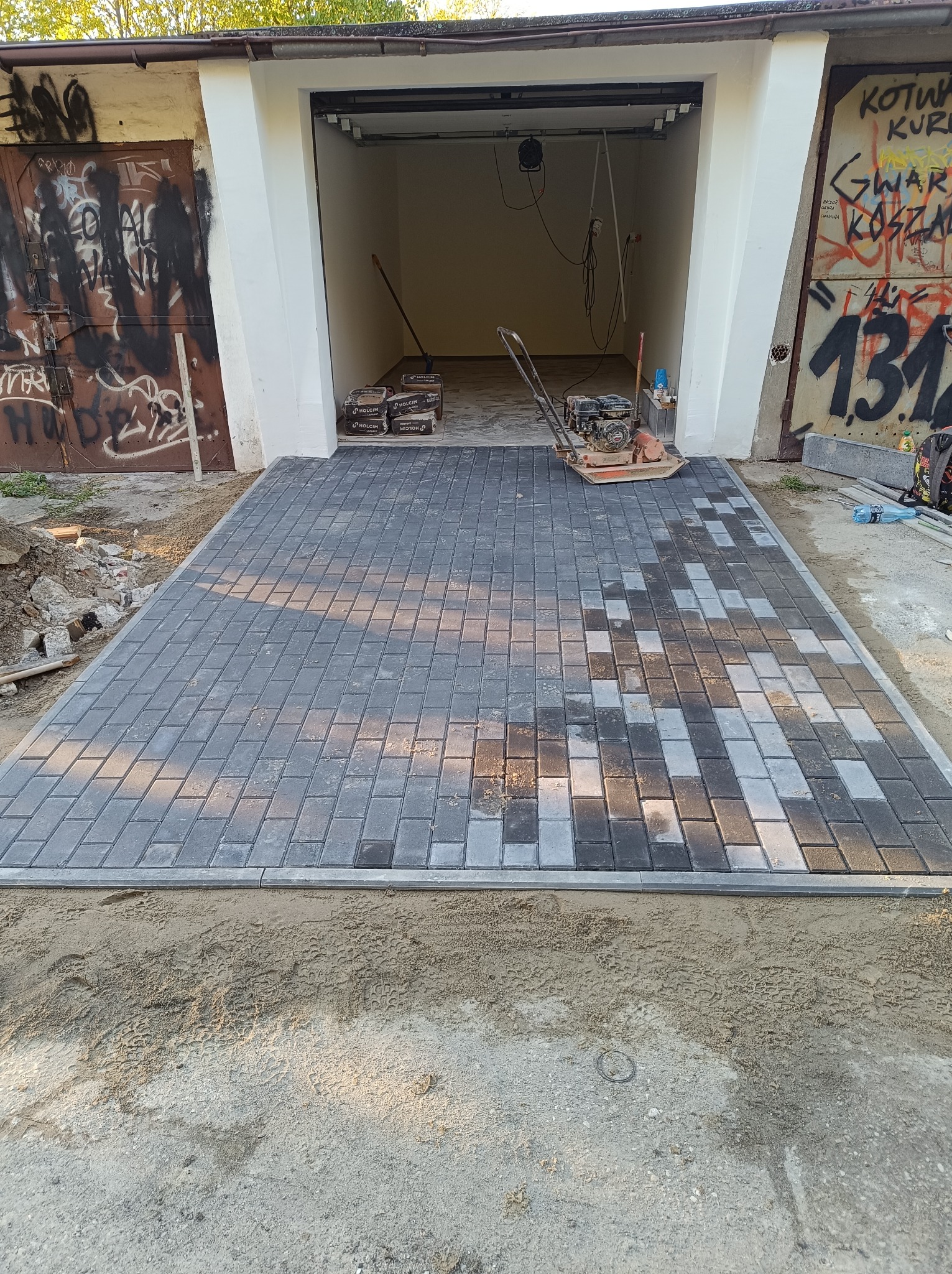 Ułożona kostka brukowa w odcieniach szarości prowadząca do garażu z graffiti na ścianach, widoczna ubijarka wibracyjna i worki z materiałem budowlanym wewnątrz.