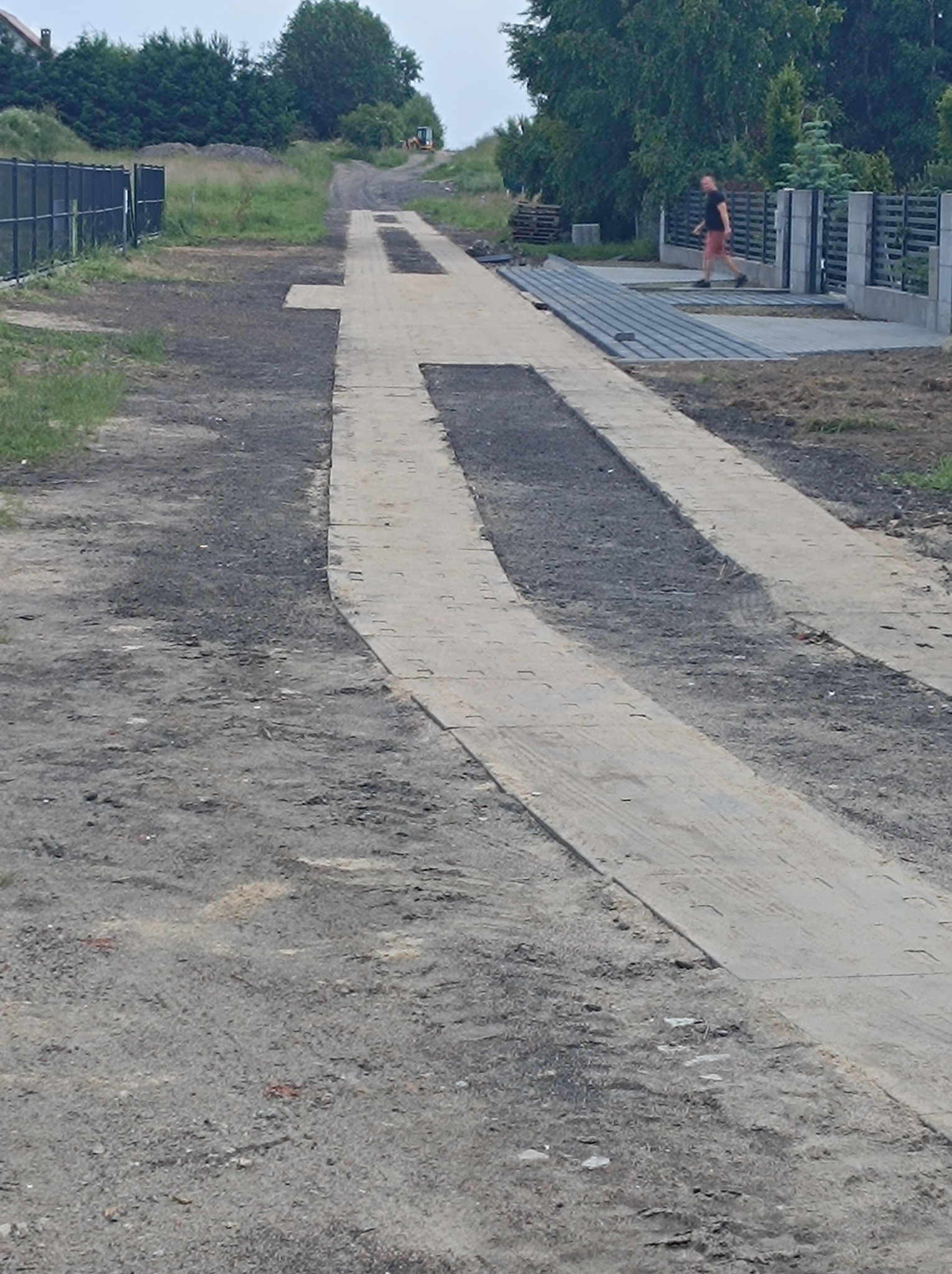 Ułożona, częściowo zasypana piaskiem, jasnoszara ścieżka z kostki prowadząca w głąb posesji, w tle koparka na wzniesieniu i spacerujący mężczyzna w czerwonych spodenkach.