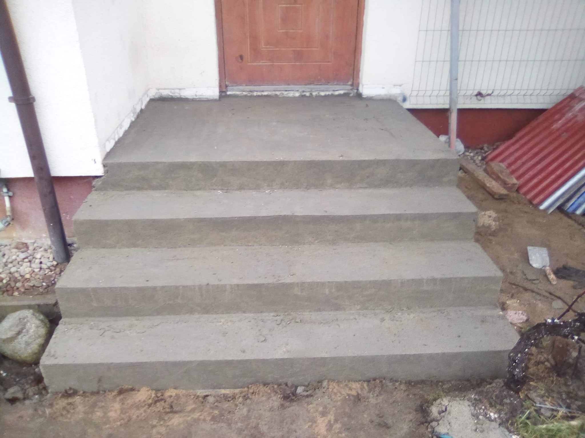 Nowe, betonowe schody wejściowe do domu, jeszcze w trakcie budowy, z widocznymi śladami zaprawy i narzędziami na ziemi.