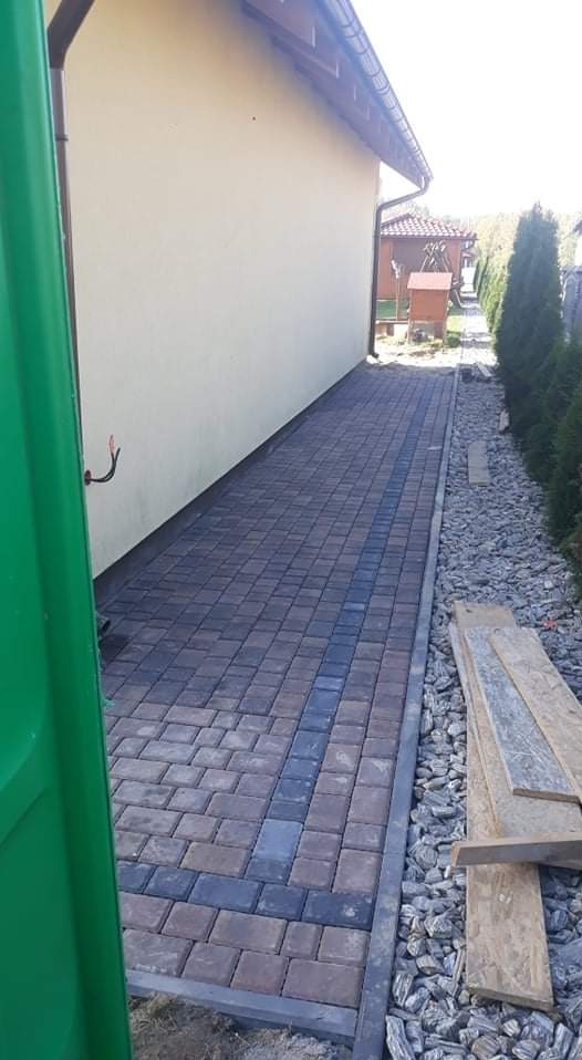 Ułożona ścieżka z kostki brukowej w odcieniach szarości i brązu, z ciemniejszym pasem pośrodku, obok ściany budynku i rabaty z kamieniami oraz deskami.