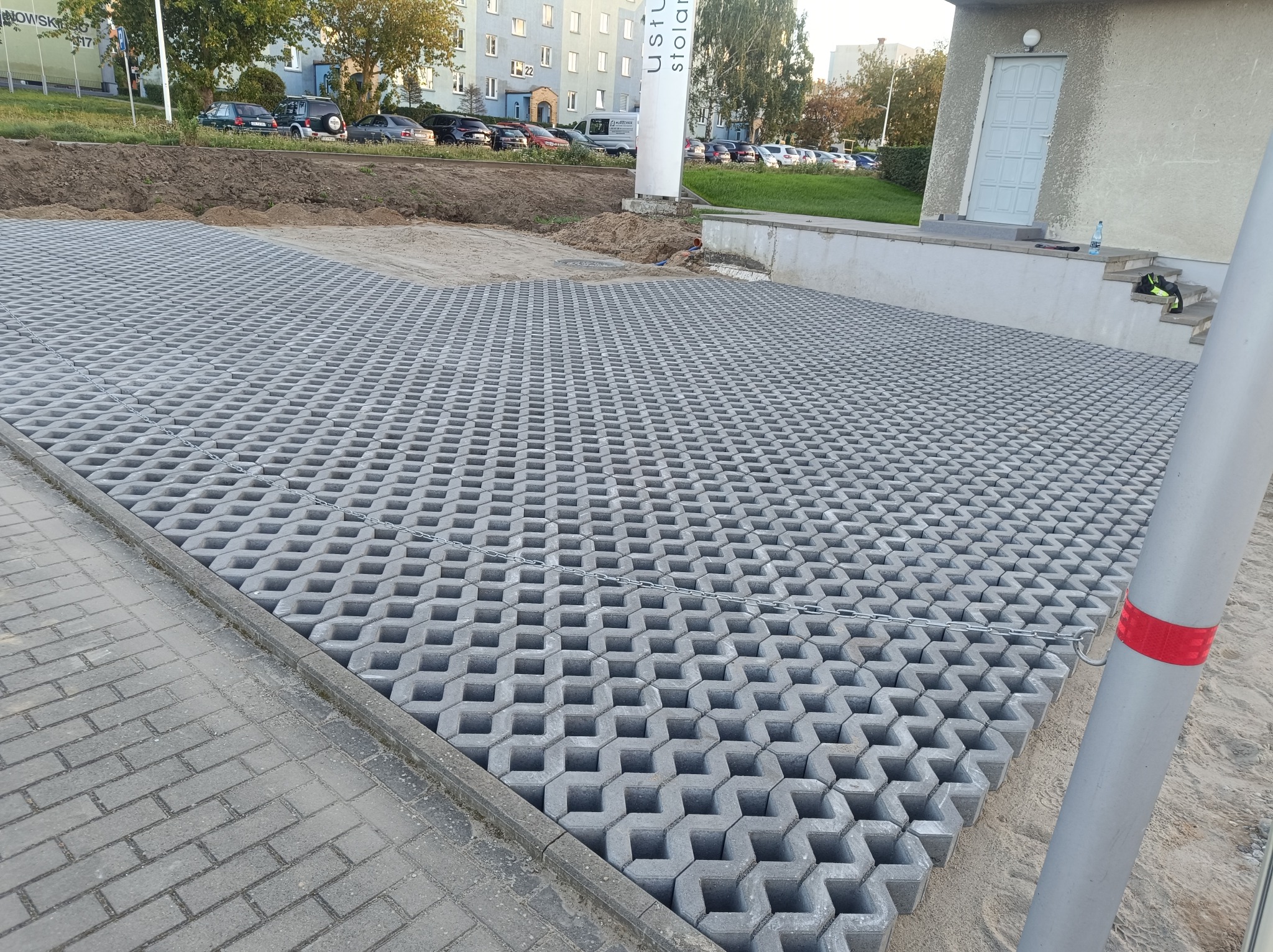 Ułożona szara ażurowa kostka brukowa na przygotowanym terenie, widoczna krawędź chodnika z kostki brukowej i budynek w tle.