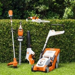 Tomas ogrody - Zestaw akumulatorowych narzędzi ogrodniczych marki Stihl: kosiarka, nożyce do żywopłotu, dmuchawa, podkaszarka i pilarka łańcuchowa, ustawione na trawie przed zielonym żywopłotem.
