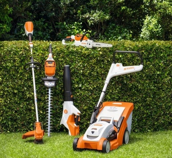 Zestaw akumulatorowych narzędzi ogrodniczych marki Stihl: kosiarka, nożyce do żywopłotu, dmuchawa, podkaszarka i pilarka łańcuchowa, ustawione na trawie przed zielonym żywopłotem.
