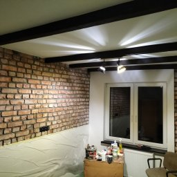 Len-Bud - Pokój w trakcie prac wykończeniowych: ściana z cegły częściowo zabezpieczona folią malarską, drewniane belki na suficie z reflektorami, okno z parapetem, grzejnik i narzędzia malarskie na skrzynce.