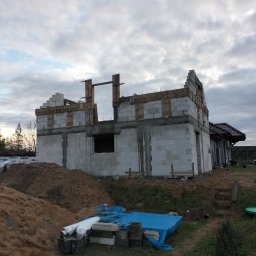 Len-Bud - Budowa domu jednorodzinnego w trakcie wznoszenia ścian z bloczków betonowych, widoczne szalunki i stropy, w tle gotowy budynek z ciemnym dachem.