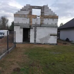 Surowy stan budynku w trakcie budowy z białych bloczków, widoczne drewniane szalunki wokół otworów okiennych i drzwiowych, częściowo ogrodzony teren budowy z widocznym samochodem dostawczym w tle.