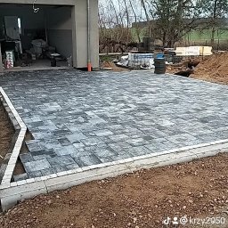 Len-Bud - Układanie szarej kostki brukowej na podjeździe przed garażem, widoczne palety z kostką i przygotowane podłoże.