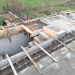 Len-Bud - Konstrukcja z betonu i zbrojenia w trakcie budowy, widoczne drewniane szalunki i elementy stalowe, w tle ogrodzenie siatkowe i pole uprawne.