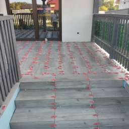 Len-Bud - Układanie szarych płytek imitujących drewno na schodach i tarasie z użyciem czerwonych klinów poziomujących, widoczny kubek na tarasie i częściowo odbicie osoby w szklanych drzwiach.