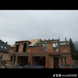 Len-Bud - Surowy stan budynku z czerwonej cegły z częściowo wykonanym dachem z drewnianych desek, widoczny szkielet konstrukcji i fragment dachu sąsiedniego budynku, pochmurne niebo w tle.