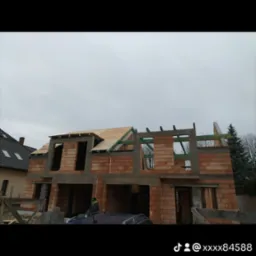 Surowy stan budynku z czerwonej cegły z częściowo wykonanym dachem z drewnianych desek, widoczny szkielet konstrukcji i fragment dachu sąsiedniego budynku, pochmurne niebo w tle.