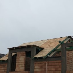 Len-Bud - Surowa konstrukcja dachu z deskowaniem na ceglanym domu w trakcie budowy, widoczne elementy betonowe i zielone krokwie, pochmurne niebo w tle.