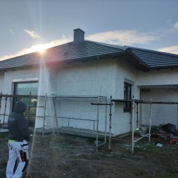 Len-Bud - Dom jednorodzinny w trakcie ocieplania, widoczne rusztowania, pracownik w roboczym ubraniu i worki z materiałem izolacyjnym, jasne niebo z promieniami słonecznymi w tle.